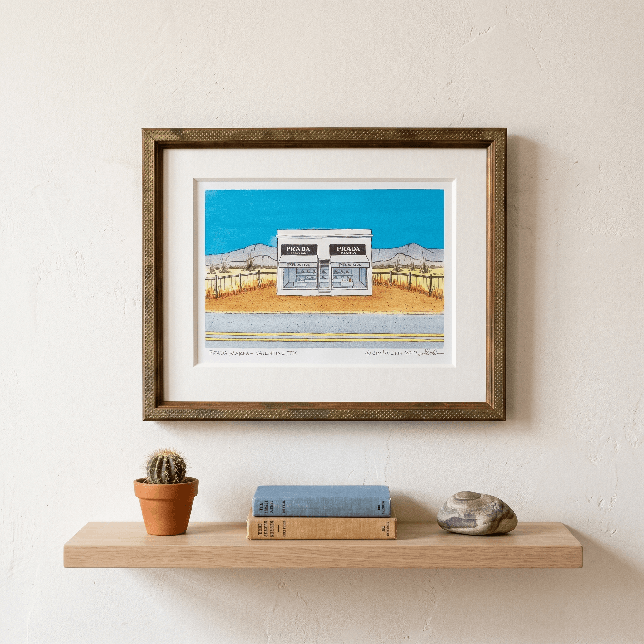 A framed Prada Marfa watercolor in a textured bronze frame on a warm wall